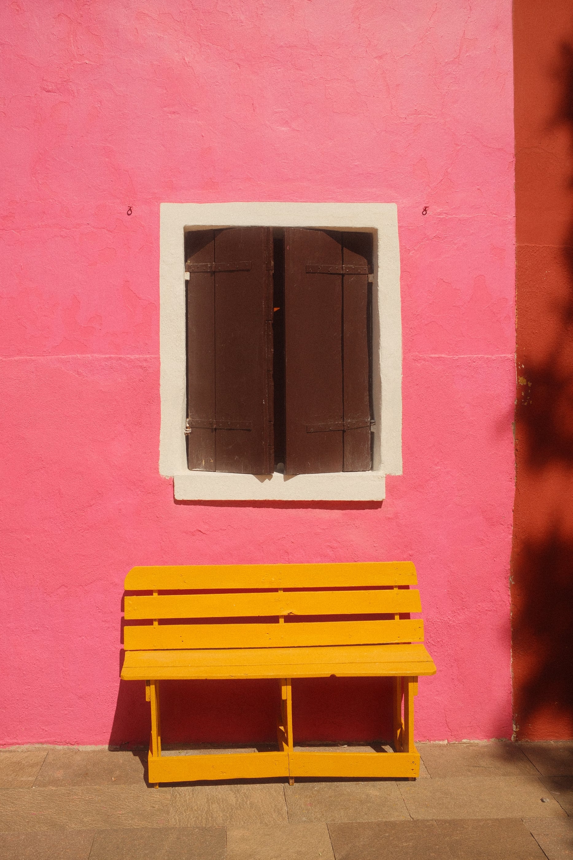 Un banc peint en jaune sous une fenêtre aux volets bruns, contre un mur rose vif.