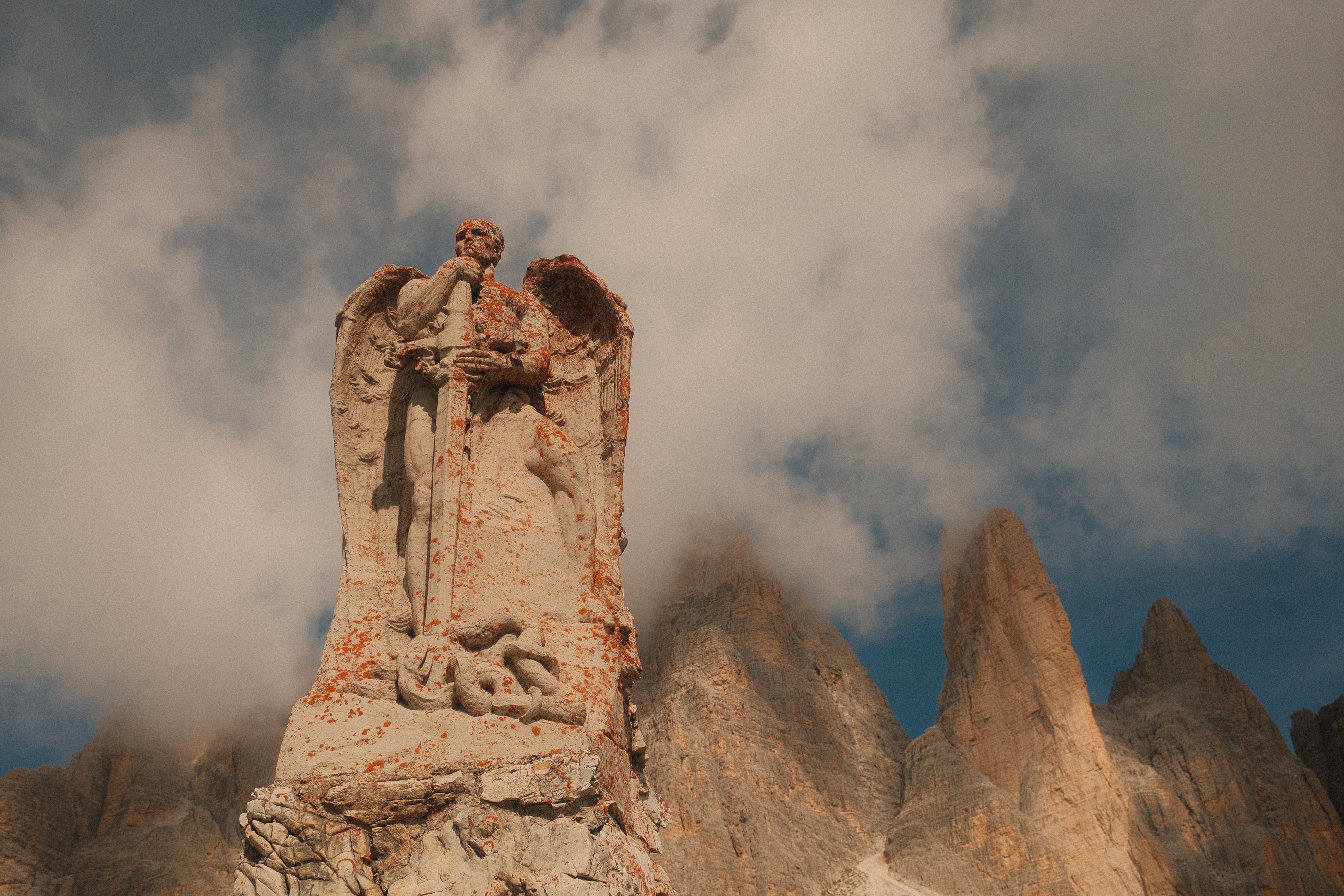 Une statue d'ange ailé appuyé sur une épée, sculptée dans la pierre, devant des pics rocheux pris dans les nuages.